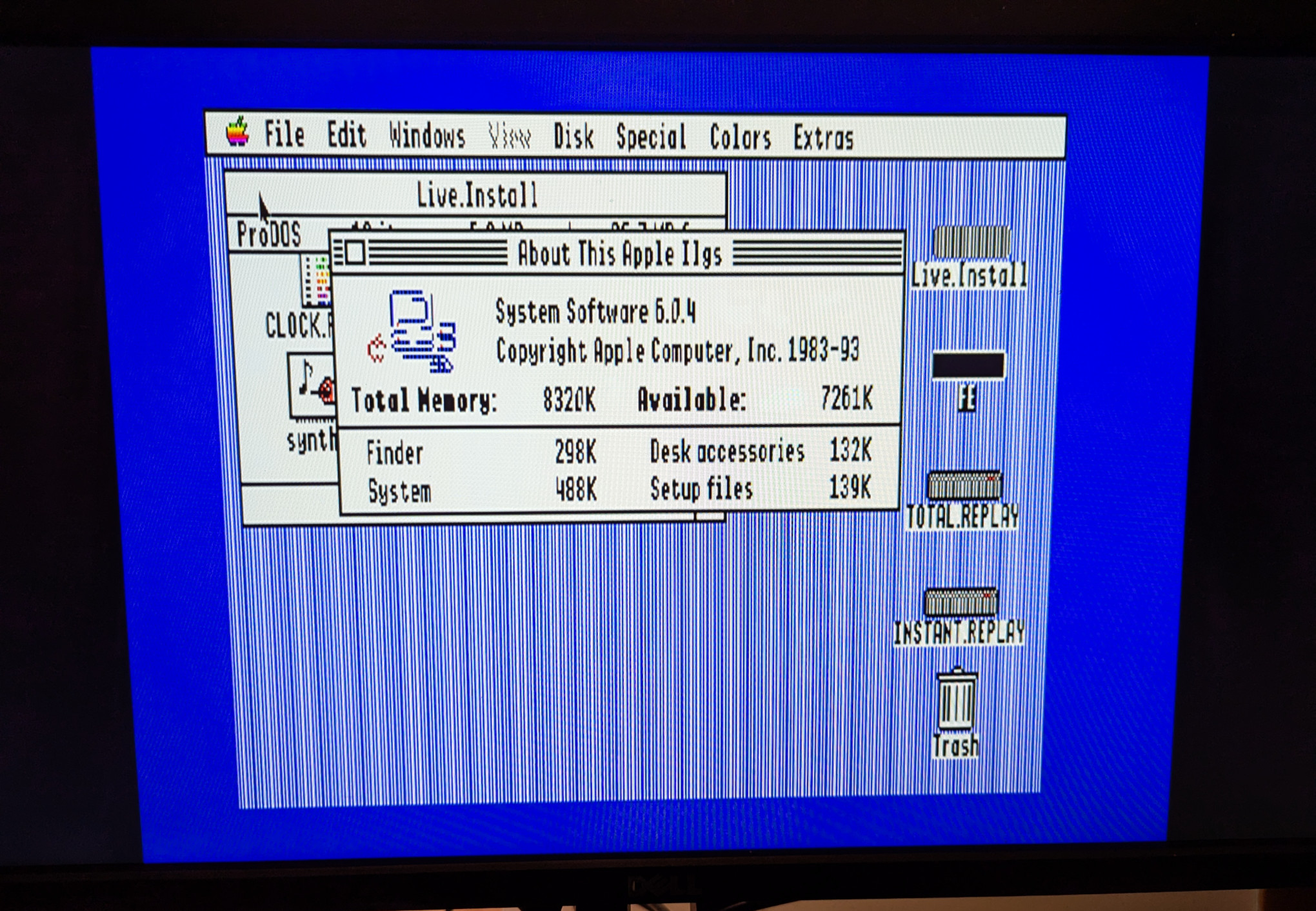 A photograph of a flat panel monitor displaying the GS/OS desktop environment running on an Apple IIgs computer.  The computer desktop is similar to a Macintosh desktop of the same era (1987), with a taskbar on the top of the screen, drive icons and a Trash can on the right, and rectangular windows for dialogs.  The window in the foreground is 'About this Apple IIgs', and shows that the computer is running System Software 6.0.4 ith 8 MiB of memory in total.  The colors are bright and it's usable, but the image is fuzzy, with edges and sharp corners looking softened as if seen through thick glass.