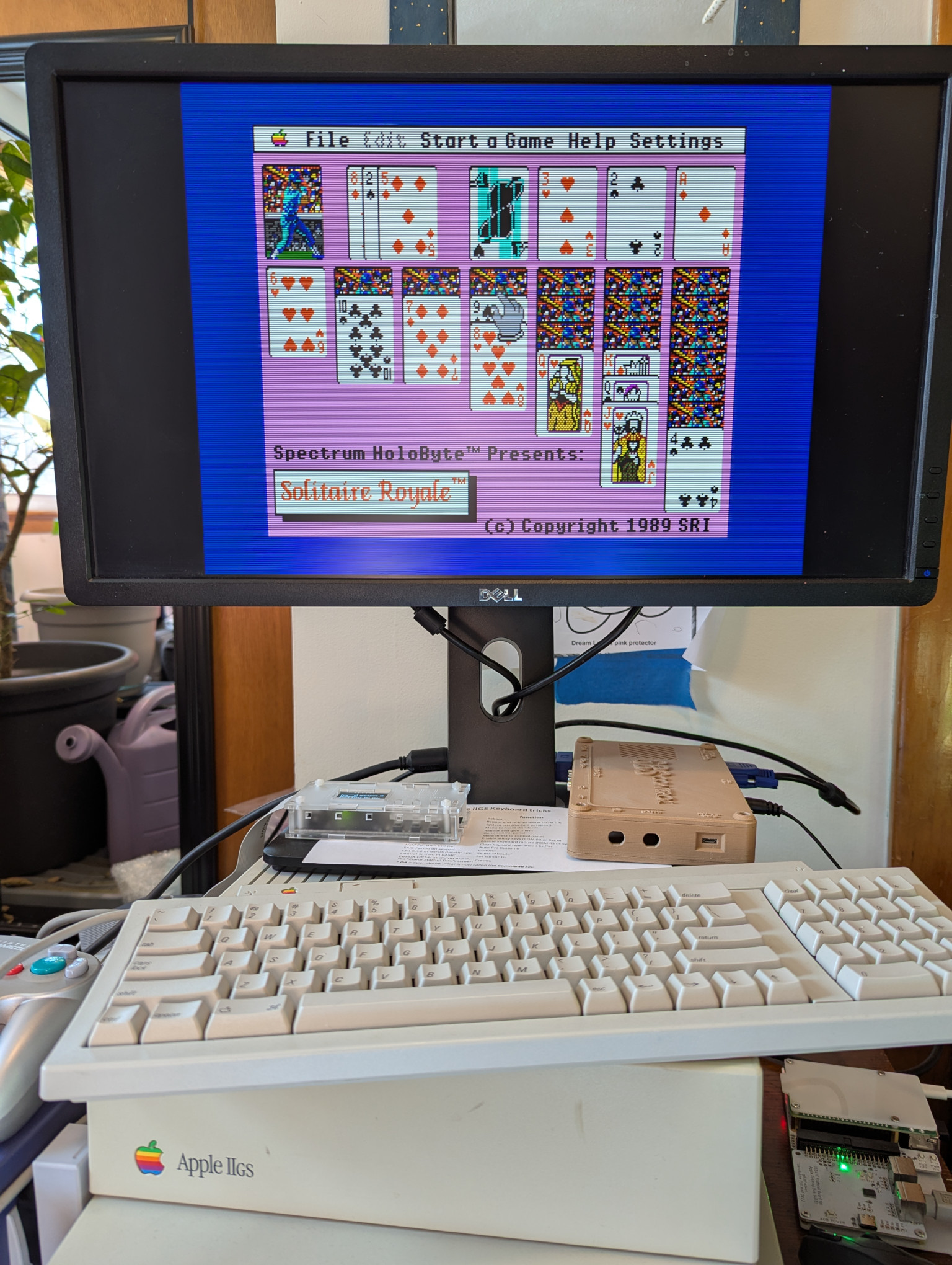 A photograph of an Apple IIgs computer, displaying the title screen from the game 'Solitaire Royale'.  The screen shows a hand of Klondike Solitaire laid out against a lavender background.  The card backs are a colorful depiction of baseball player at bat, in a photorealistic style.