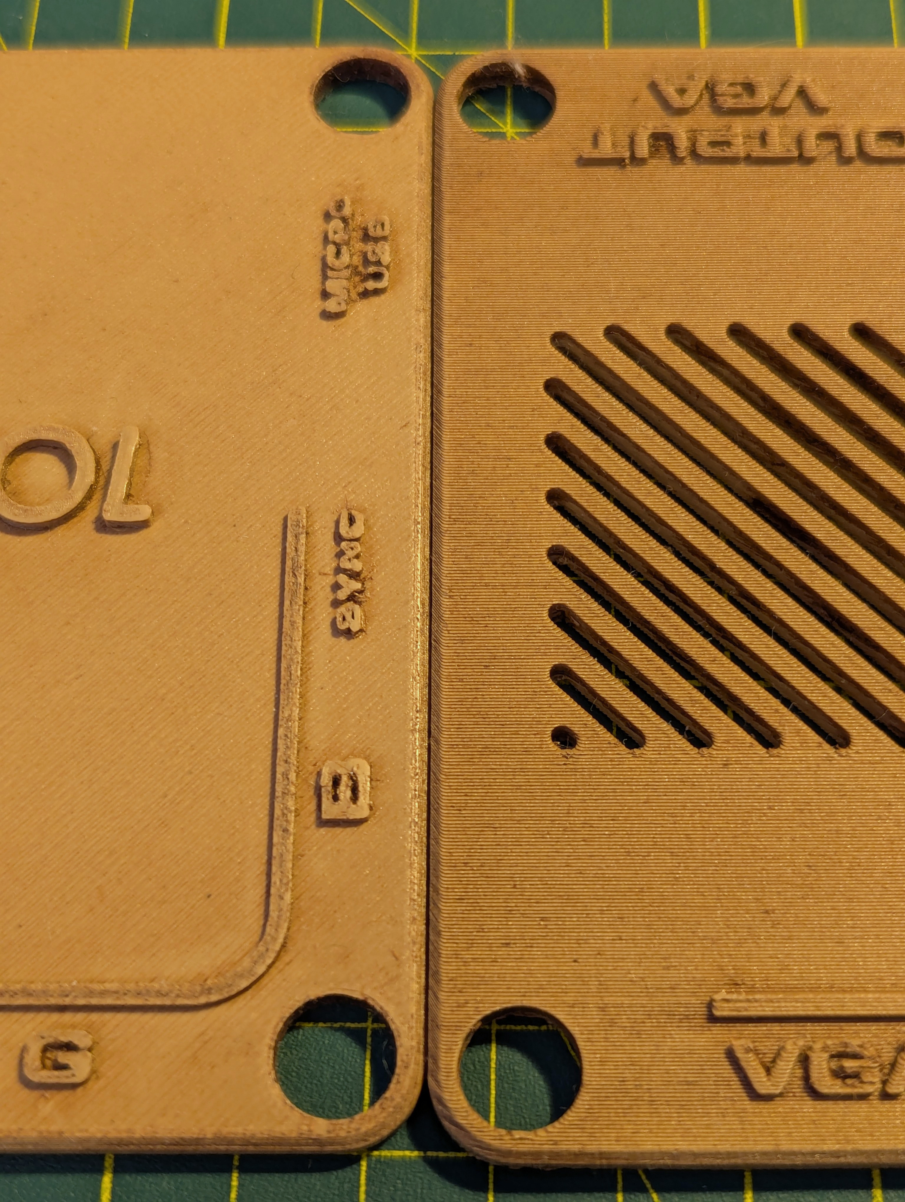 A close-up photograph with the old top plate for the GBS-C enclosure on the left, and the new top plate on the right.  Both have been stained with a wood stain.  The old plate on the left still looks like shiny tan plastic, but the one on the right has a darker coloration and 'grain lines' due to the printing method I used for it.